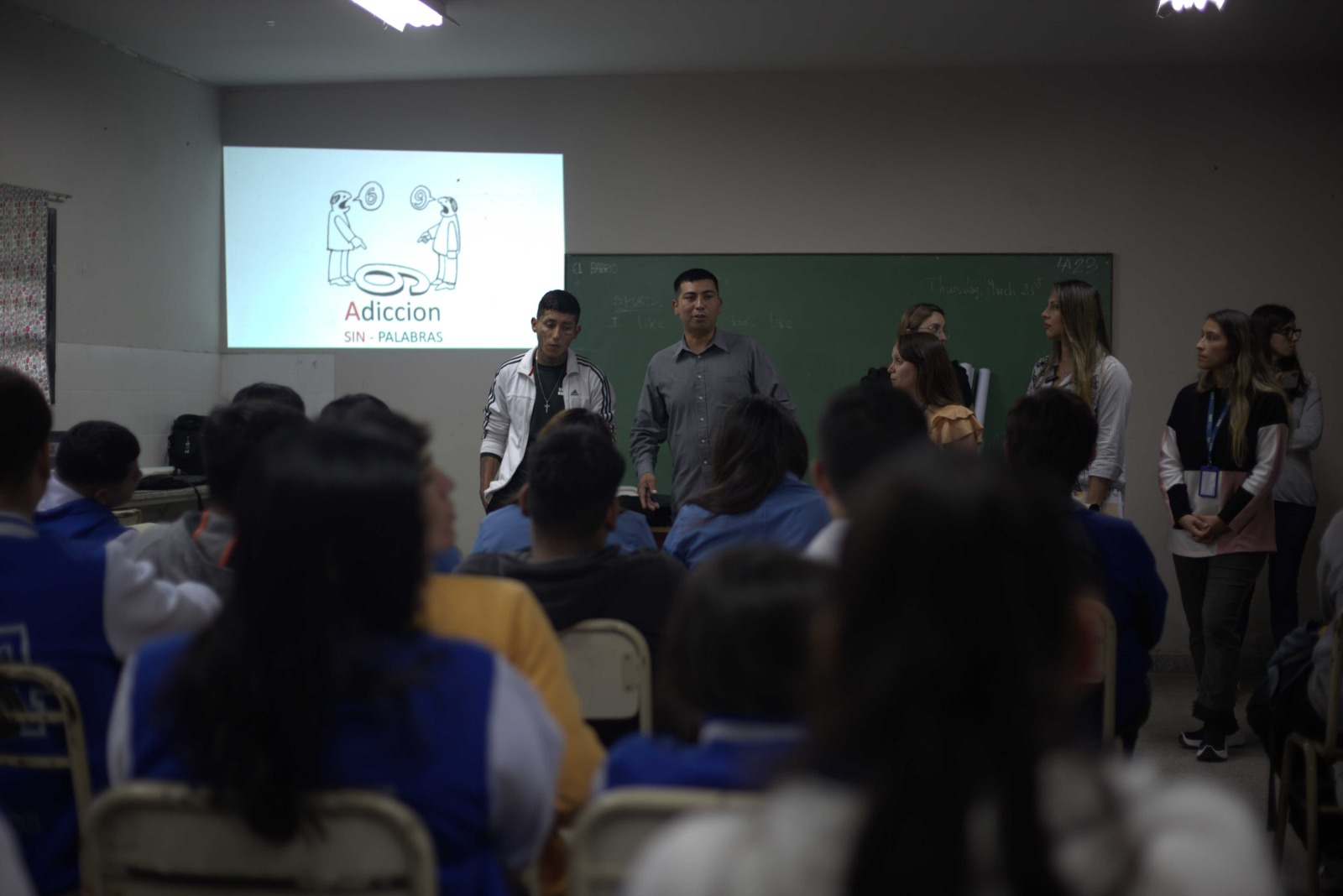 Coordinador y joven exponiendo frente a estudiantes con una proyección sobre adicciones